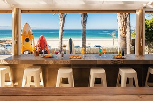 A beachside bar with a view of the ocean, featuring kayaks, a surfboard, and a variety of food and drinks on the counter.