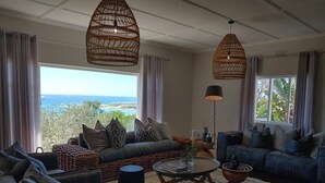 A spacious living room with a view of the sea, furnished with sofas, a coffee table, and hanging wicker lamps. - The Bungalow  (Plettenberg Bay)