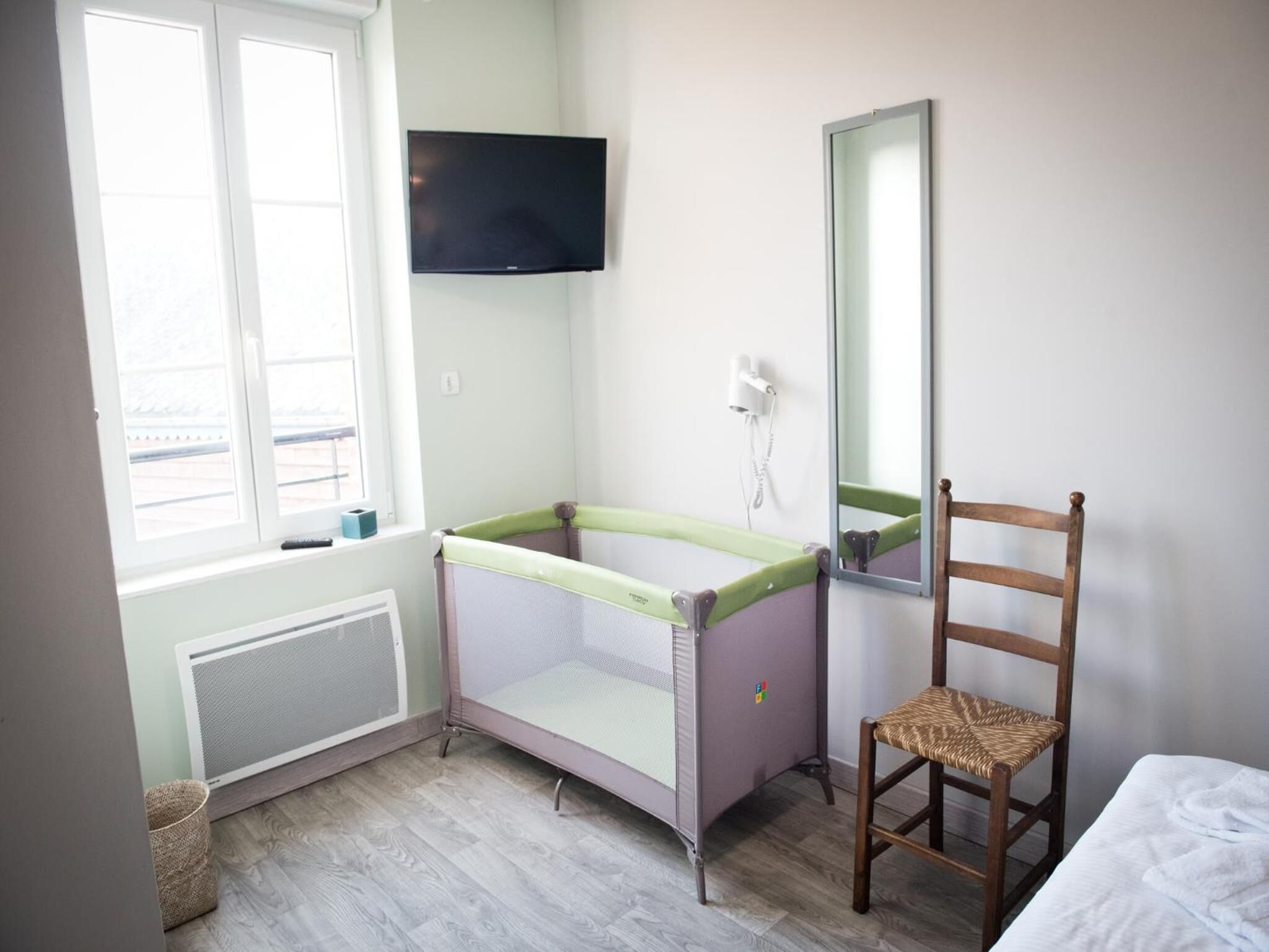 A small, well-lit room with a window, a television mounted on the wall, a chair, and a bed.