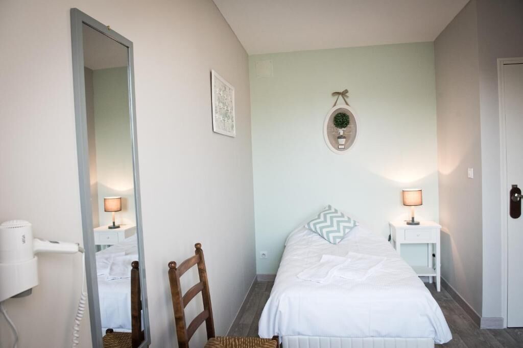 Quarto com cama, uma cadeira, um espelho, luminária na parede e uma pequena mesa com abajur.