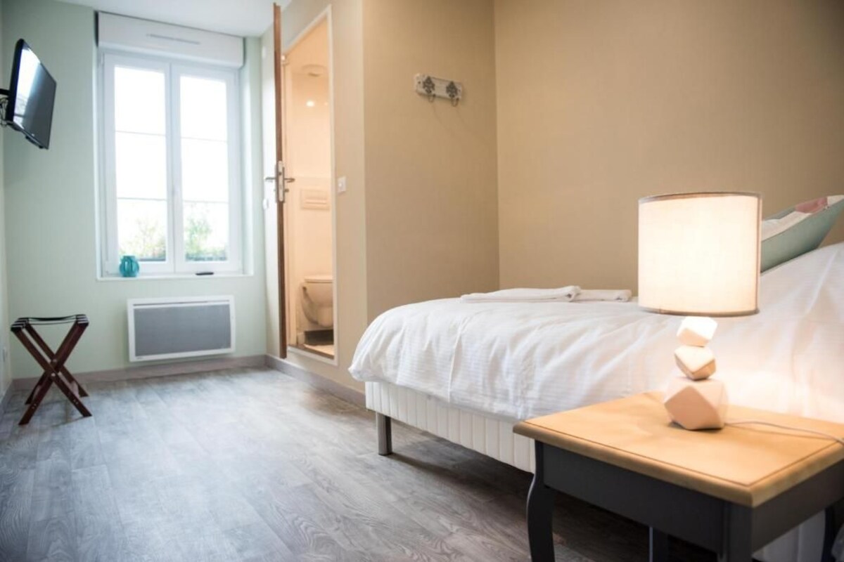 A bedroom with a bed, bedside table, lamp, TV, window, radiator, and a door leading to another room.