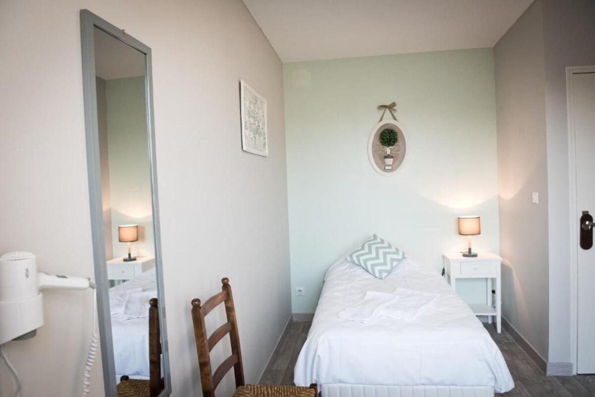 A bedroom with a bed, a chair, a mirror, a wall-mounted light fixture, and a small table with a lamp.