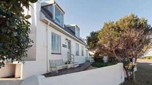 A two-story house with a white exterior, a small porch with a bench, and a tree in the front yard. - Pikkewyntjie Cottage (Hermanus)
