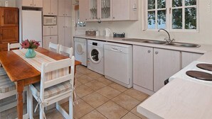 A compact kitchen with a wooden dining table, white chairs, a washing machine, and a sink. - Pikkewyntjie Cottage (Hermanus)