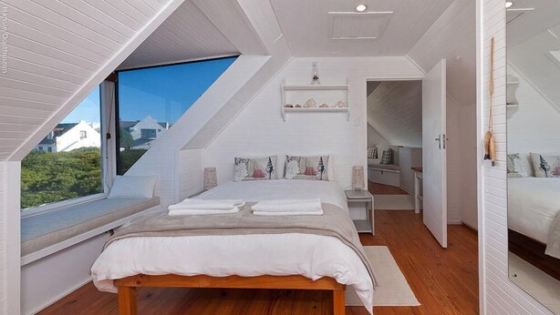 A bedroom with a large window, a bed with white linens, a wooden floor, and a small bedside table. - Pikkewyntjie Cottage (Hermanus)