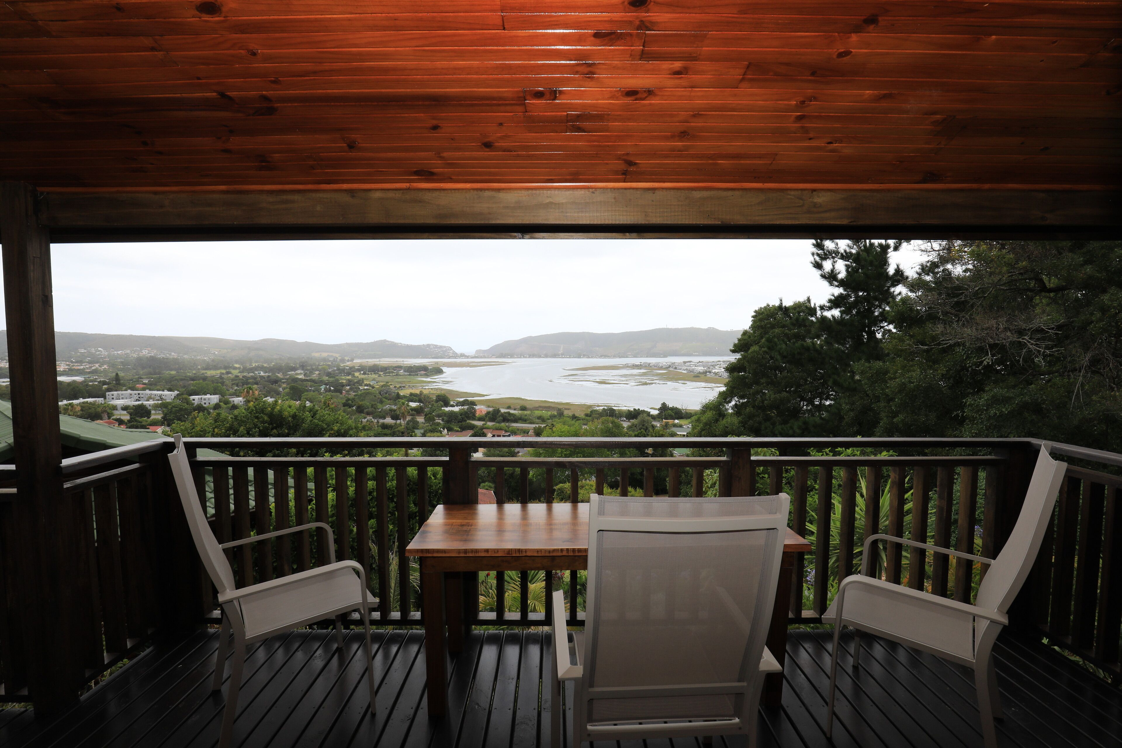 A wooden deck with a table and two chairs, overlooking a river and hills.