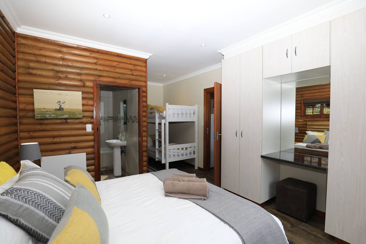 A bedroom with a bed, bedside table, and a mirror. There is a bathroom with a sink and a shower visible through an open door.