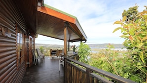 Eine Holzterrasse mit überdachtem Bereich, Sitzgelegenheiten im Freien und Blick auf einen See und Hügel.