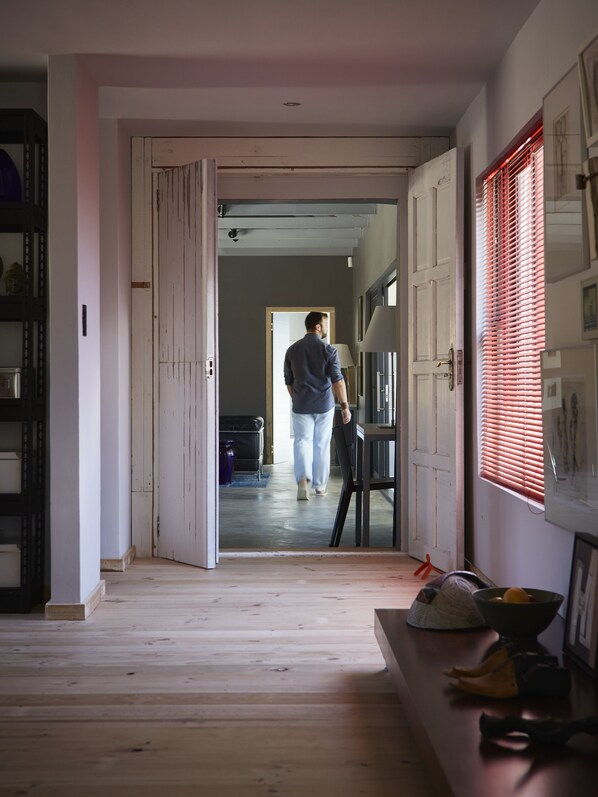 Un couloir avec un plancher en bois, une personne qui marche vers une pièce, et une fenêtre avec des stores.