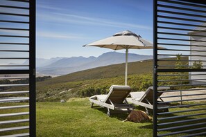 Une vue à travers une fenêtre dont le cadre est en métal, avec en premier plan une chaise longue et un parasol, un chien, et en arrière-plan un paysage montagneux.