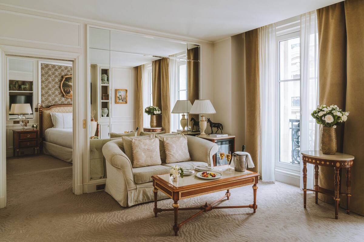 A spacious living room with a sofa, a coffee table, and a dining area with a table and chairs. There is a large mirror on the wall, a window with curtains, and a vase with flowers on a side table.