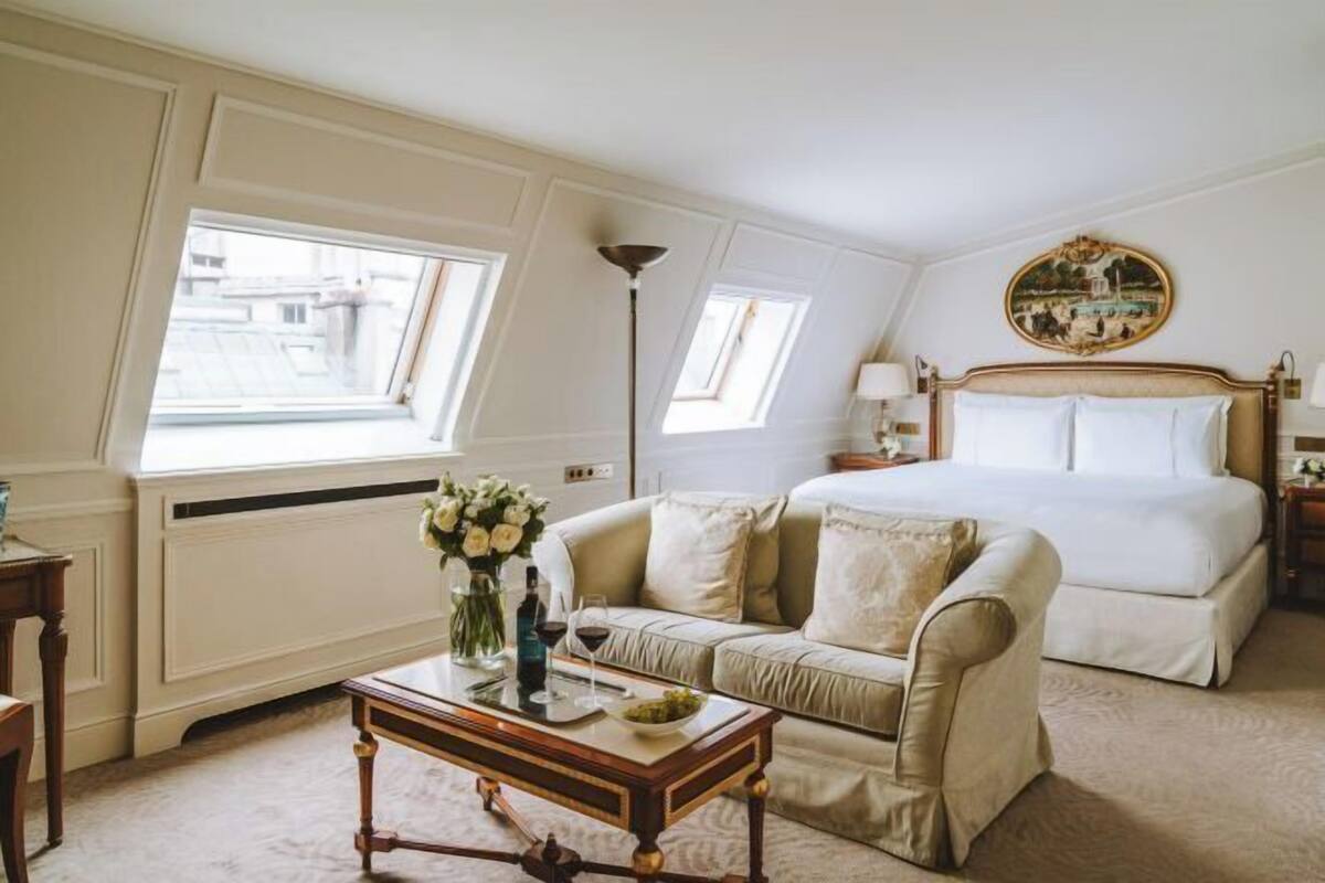 A bedroom with a bed, a sofa, a table with a vase and wine glasses, and a painting on the wall.