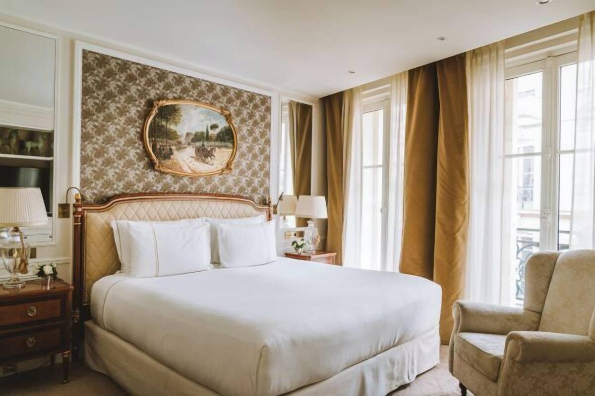 A hotel room with a large bed, a nightstand, a chair, a painting on the wall, and a window with curtains.