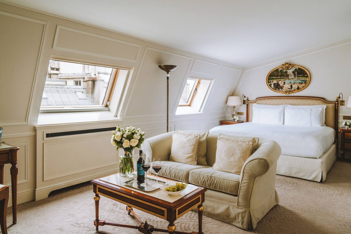 A hotel room with a large bed, a sofa, a coffee table with flowers, and a view of buildings through a window.