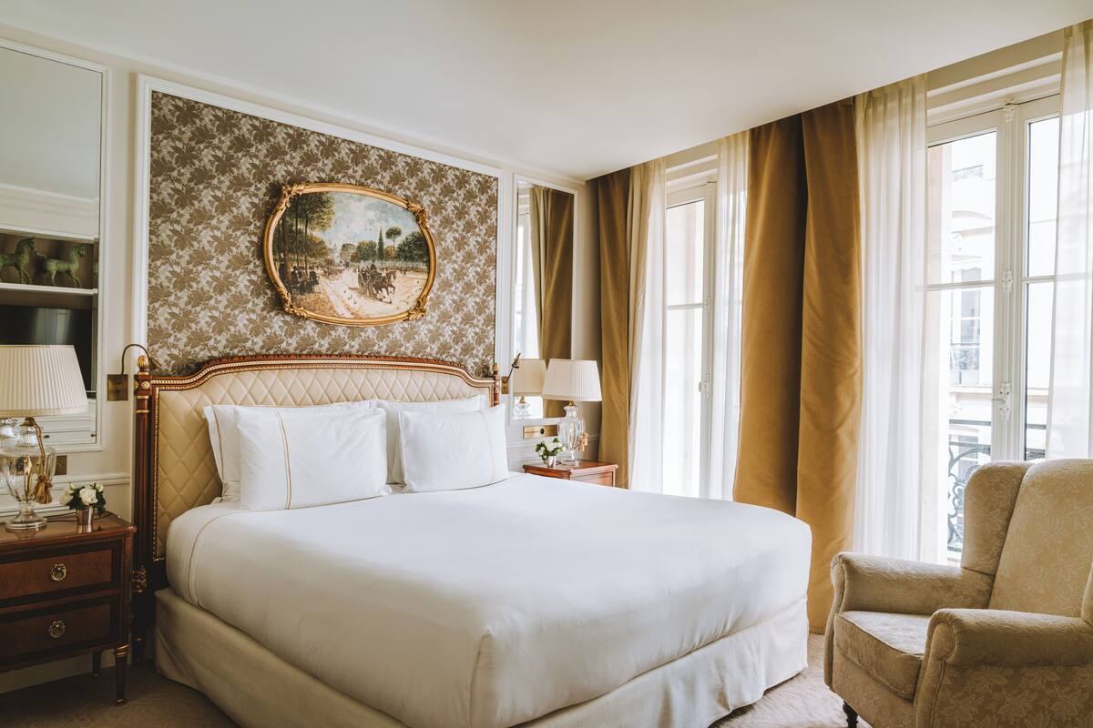 A large bed with a quilted headboard, a nightstand with a lamp, a chair, and a painting on the wall.