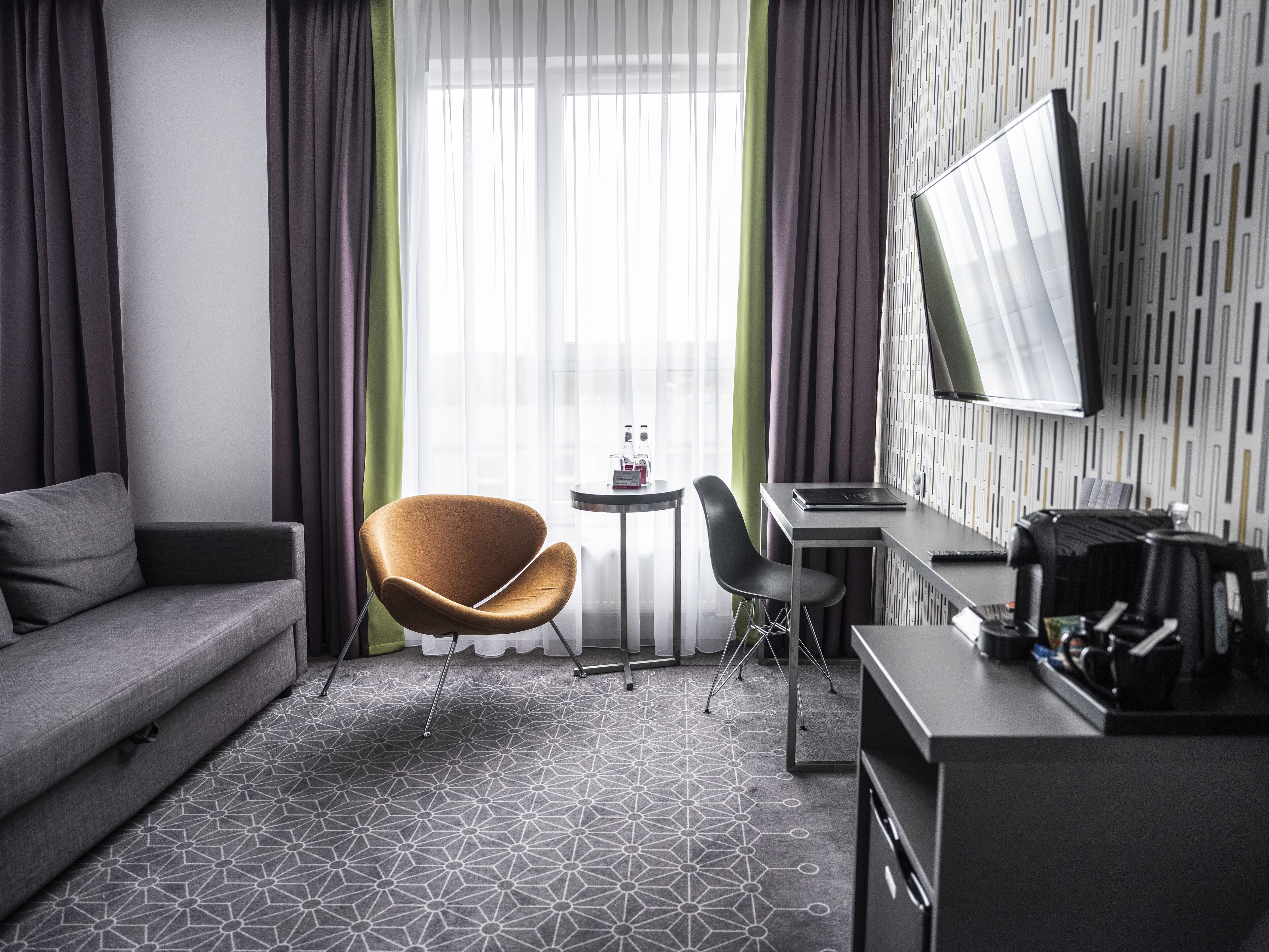 A modern hotel room with a grey sofa, a unique orange chair, a small round table with a bottle, a desk with a laptop, and a flat-screen TV mounted on the wall.