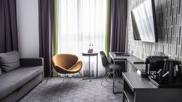 Un hotel moderno con un sofá gris, un sillón naranja único, una mesita redonda con una botella, un escritorio con una computadora portátil y un televisor de pantalla plana montado en la pared.