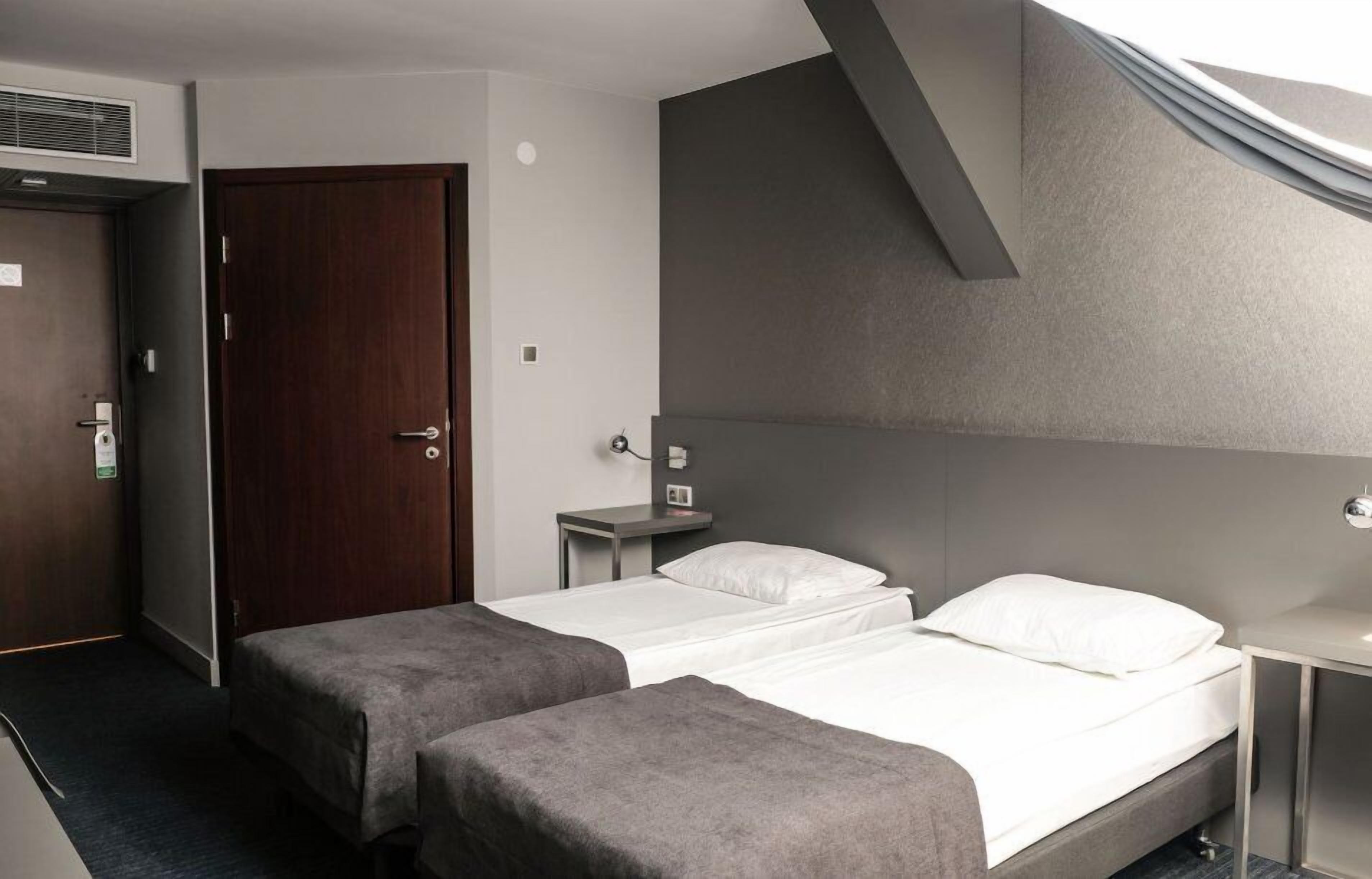 A hotel room with two single beds, a wooden door, a small table with a lamp, and a ceiling vent.