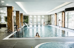 Uma piscina interna com pilares de madeira, paredes de vidro transparente e chão revestido de azulejos.
