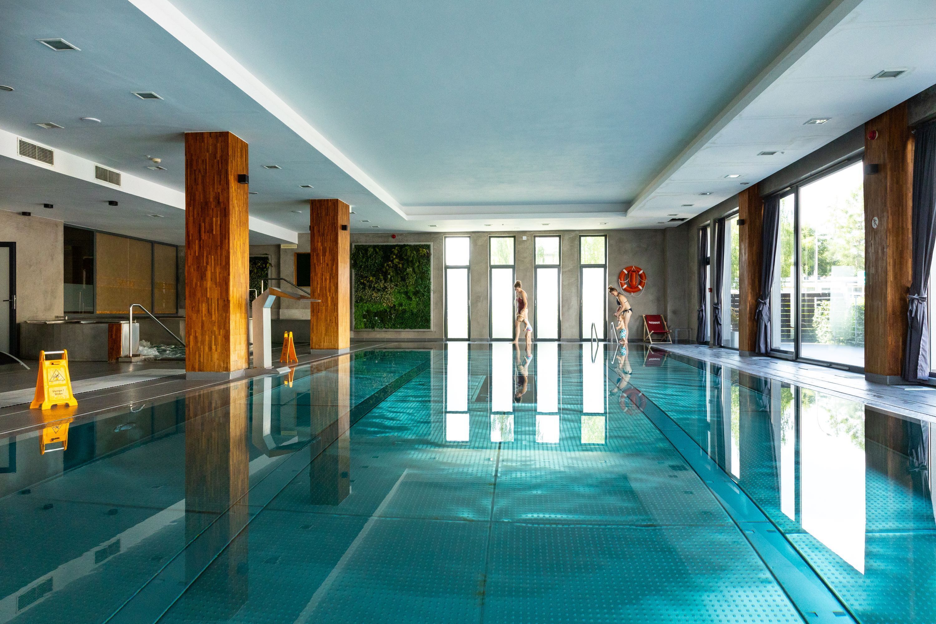 An indoor swimming pool with clear water, wooden pillars, and large windows allowing natural light.