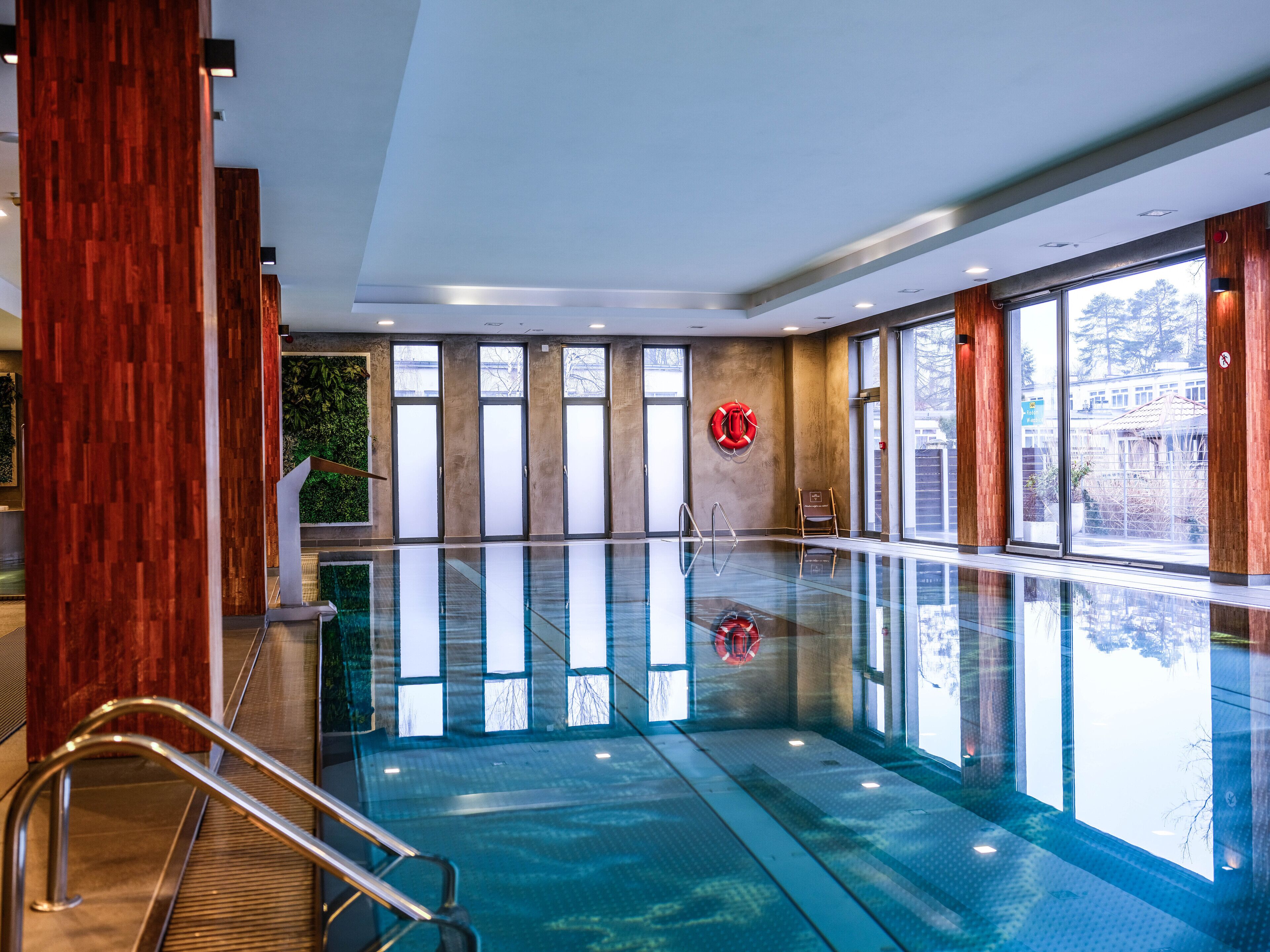 A large indoor swimming pool with clear glass walls, wooden paneling, and a lifebuoy on the wall.