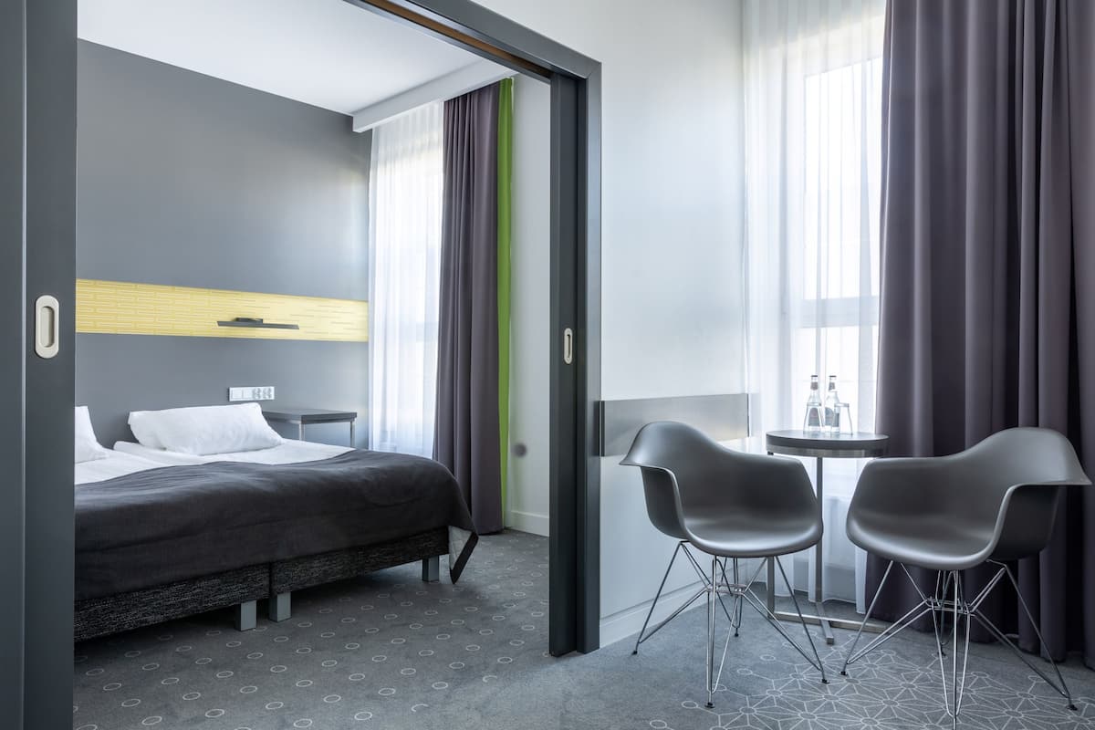 Een moderne hotelkamer met een bed, een klein tafeltje met twee stoelen en een raam met gordijnen.