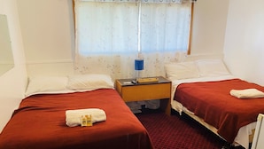 A hotel room with two beds, each with a red bedspread, white pillows, and a folded towel. There is a nightstand with a lamp, a small table, and a window with a floral curtain. - Theodore Motor Hotel (Theodore)
