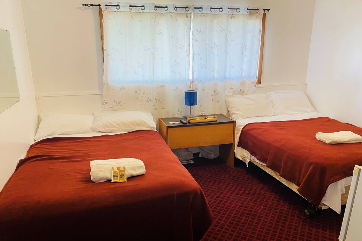 A hotel room with two beds, each with a red bedspread, white pillows, and a folded towel. There is a nightstand with a lamp, a small table, and a window with a floral curtain.