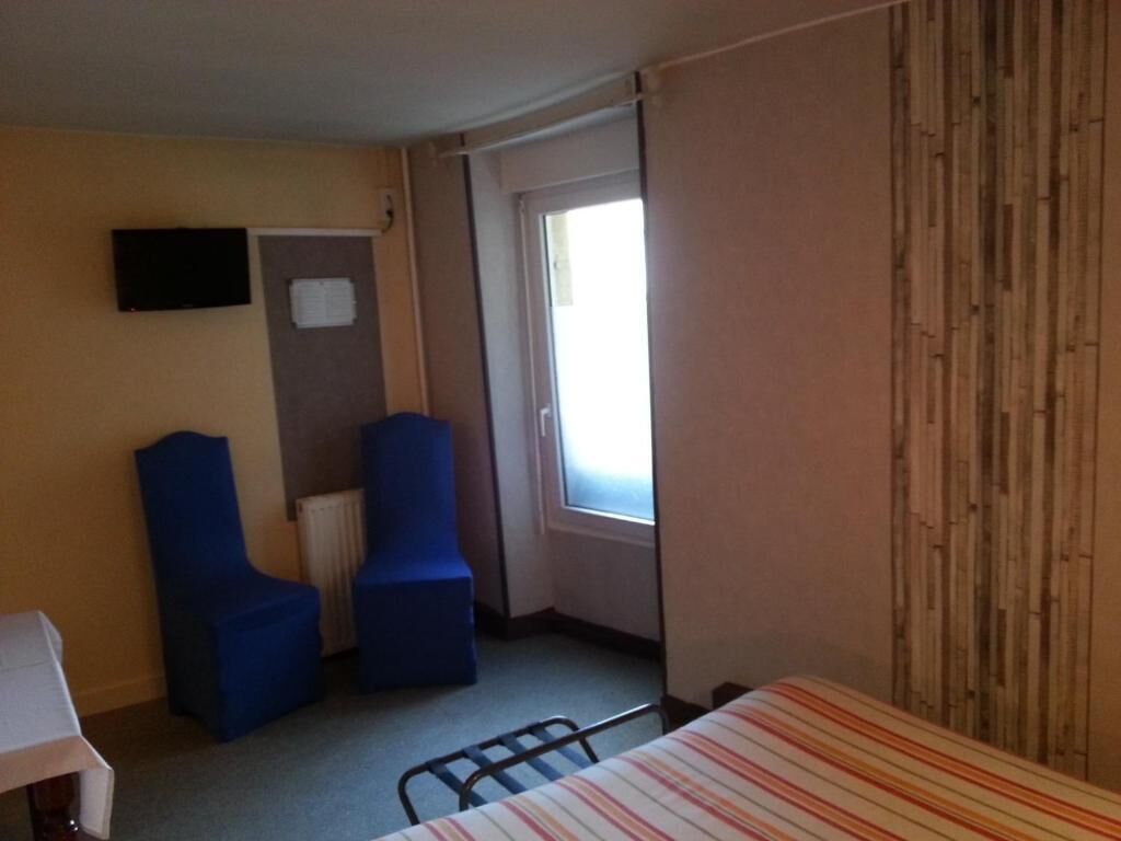 Ein kleines Hotelzimmer mit einem Einzelbett, zwei blauen Stühlen, einem kleinen Tisch, einem Fernseher und einem Fenster mit Vorhängen.