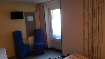 Una habitación de hotel pequeña con una cama individual, dos sillas azules, una mesita, un televisor y una ventana con cortinas.