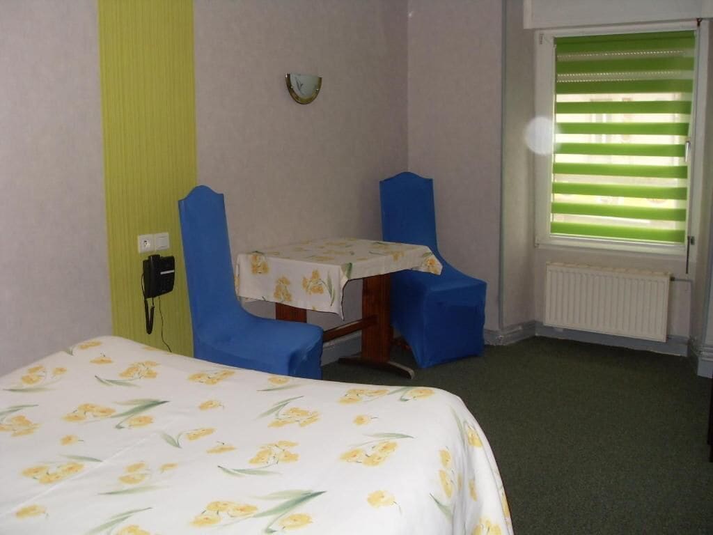 Une petite pièce comprenant un lit, une table recouverte d’une nappe, deux chaises, un radiateur et une fenêtre avec des stores.