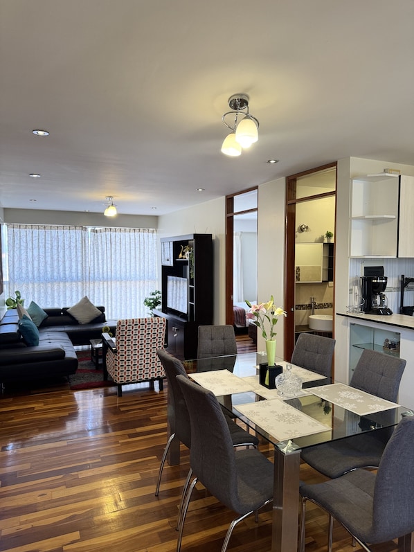 A modern living room with a sofa, dining table, chairs, and a kitchen area with cabinets and a coffee machine.