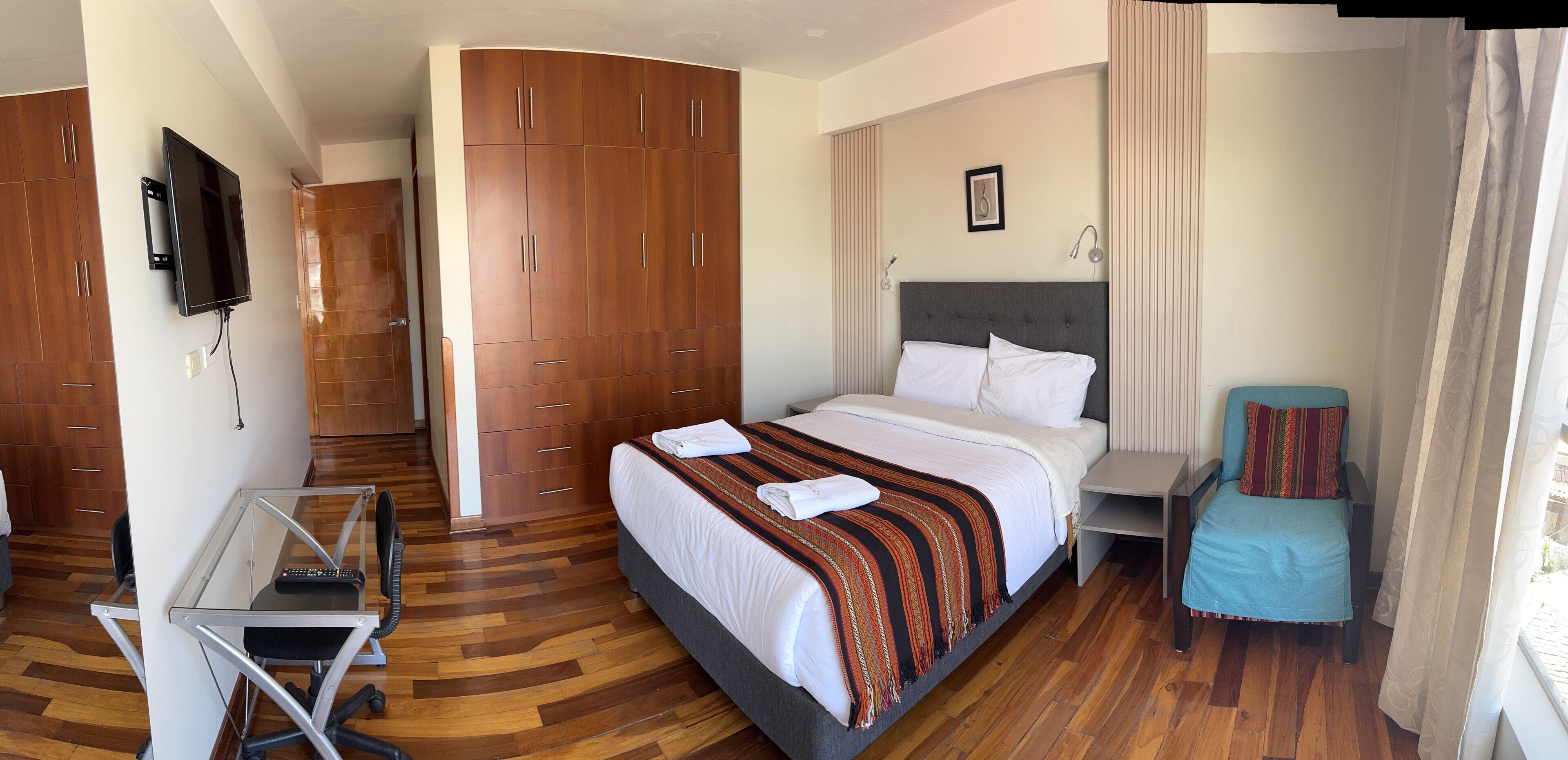 A hotel room with a bed, a desk with a chair, a TV mounted on the wall, and a wardrobe.