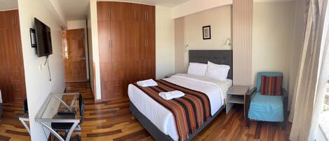A hotel room with a bed, a desk with a chair, a TV mounted on the wall, and a wardrobe.