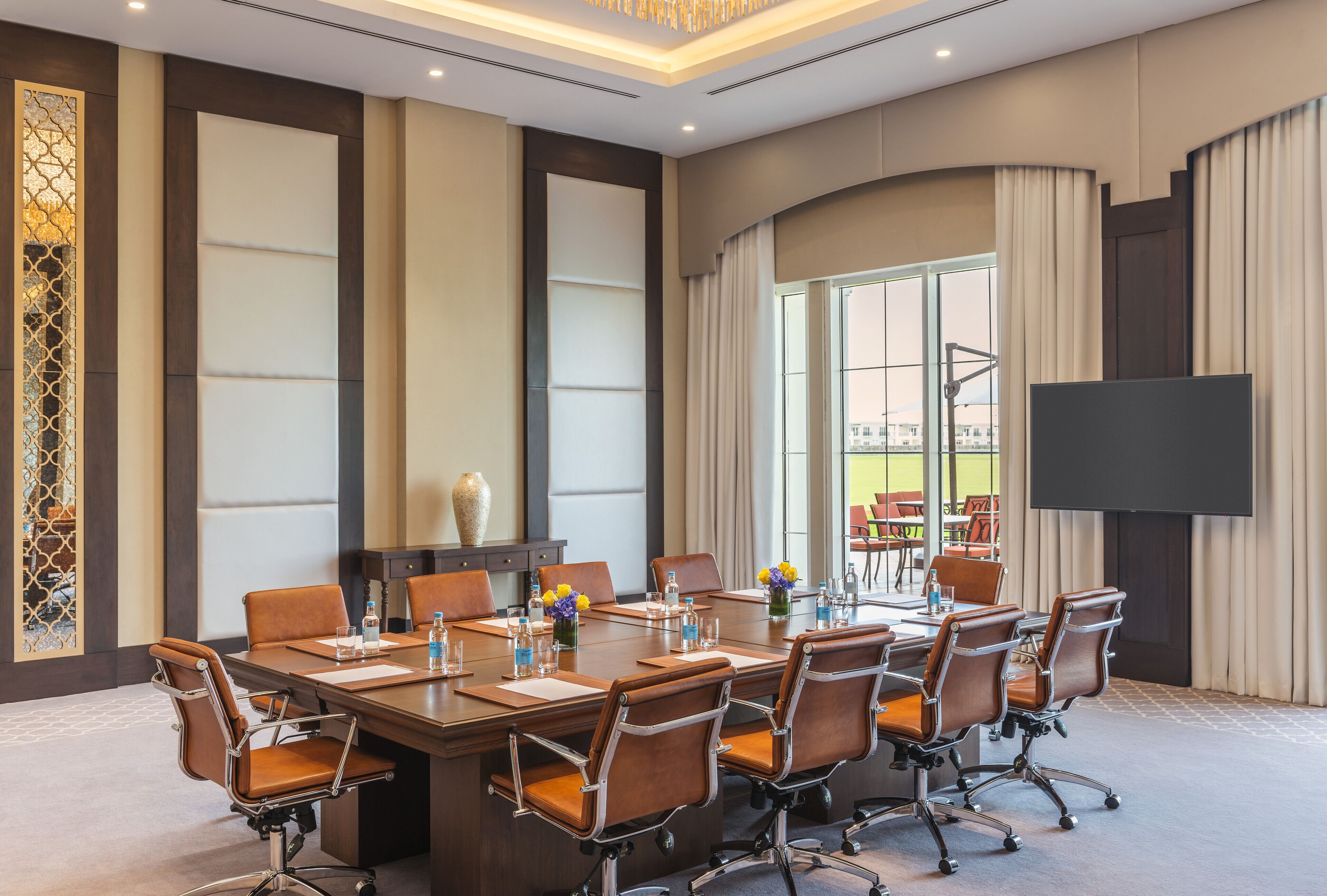 A conference room with a large table, multiple chairs, a flat-screen TV, and a decorative vase.