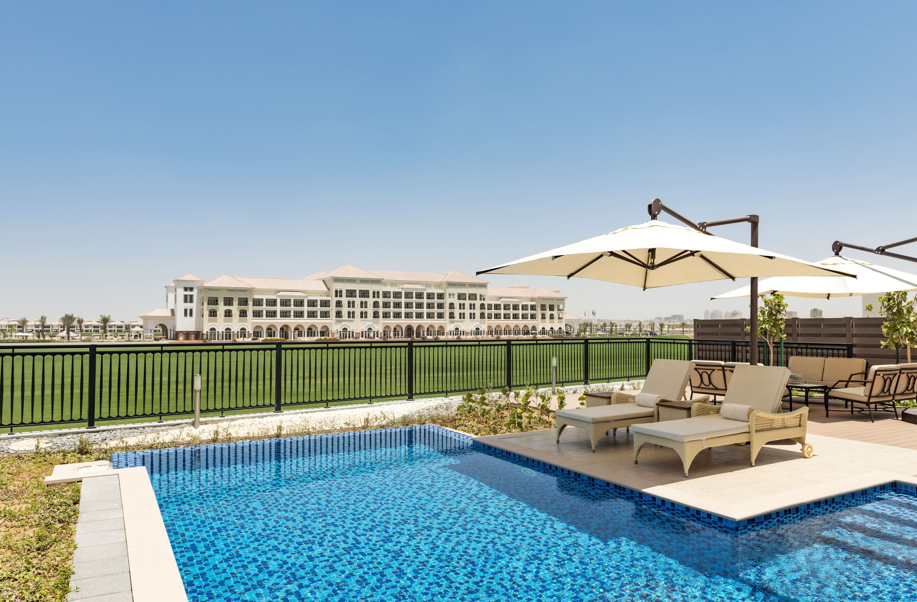 A swimming pool with lounge chairs and umbrellas, surrounded by a fence and a building in the background.