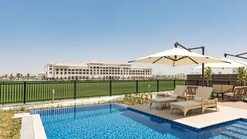 A swimming pool with lounge chairs and umbrellas, surrounded by a fence and a building in the background.