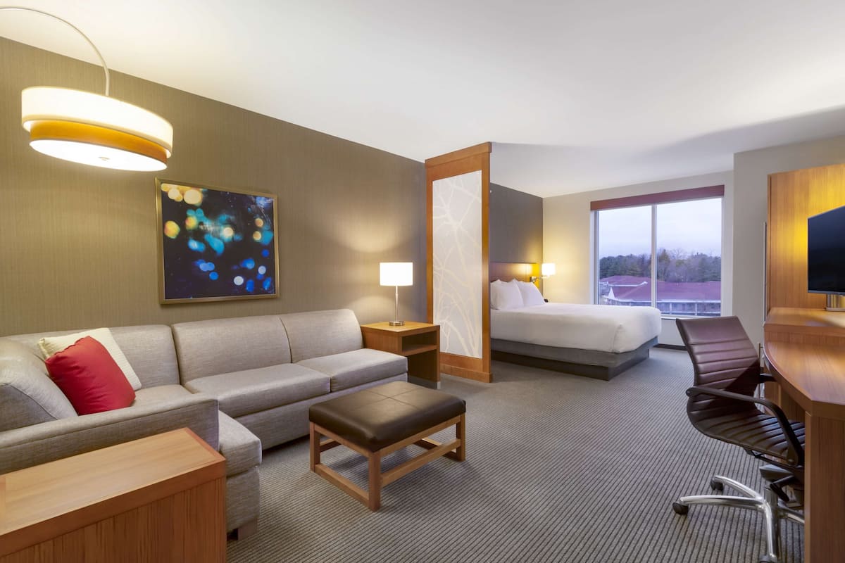 A hotel room with a sofa, a wooden coffee table, a desk with a chair, a bed, a nightstand with a lamp, and a large window offering a view.