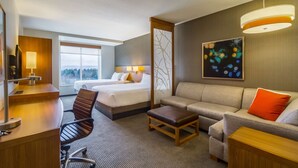 Habitación de hotel con cama, sofá, escritorio y silla. Se ve un ventanal con vistas al exterior.