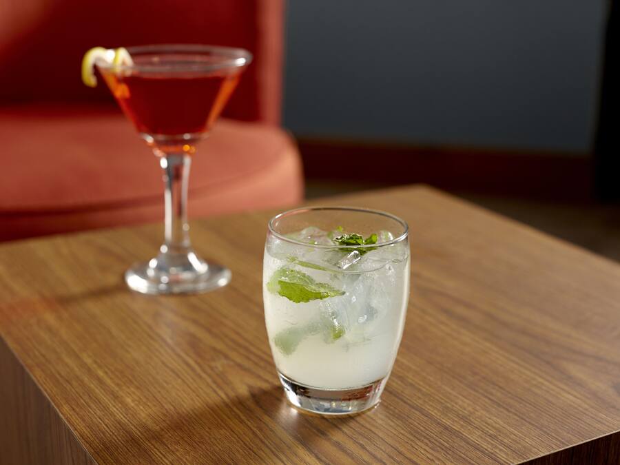 Une table en bois sur laquelle reposent deux verres contenant une boisson claire, l’un agrémenté d’une garniture, et un cocktail rouge en arrière-plan.