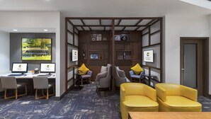 Una habitación de hotel moderna con un escritorio, dos sillas, un sofá y una pantalla de televisión que muestra el logotipo del hotel.