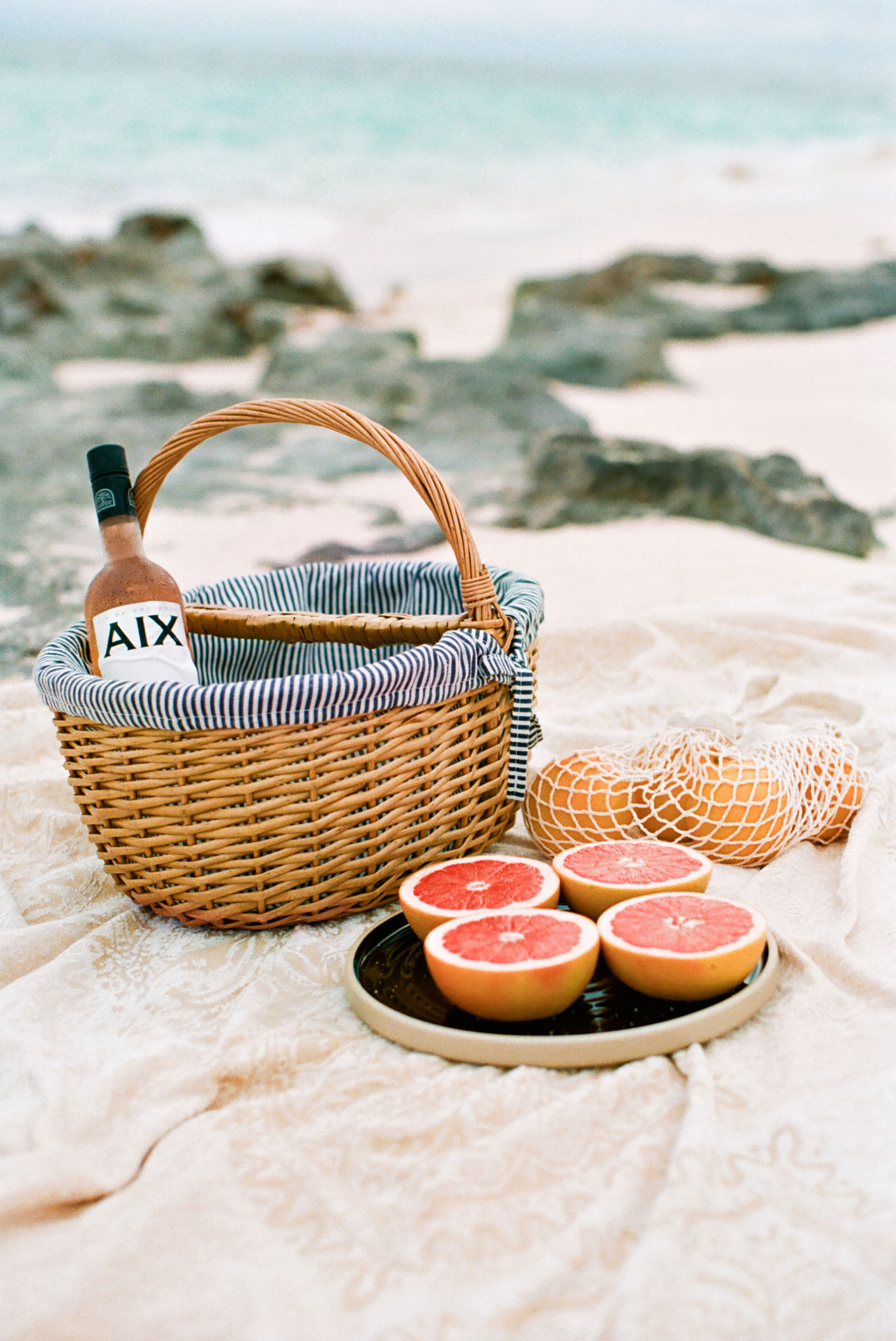 A wicker basket with a bottle labeled "AIX" and a net bag with citrus fruits on a beach blanket.
