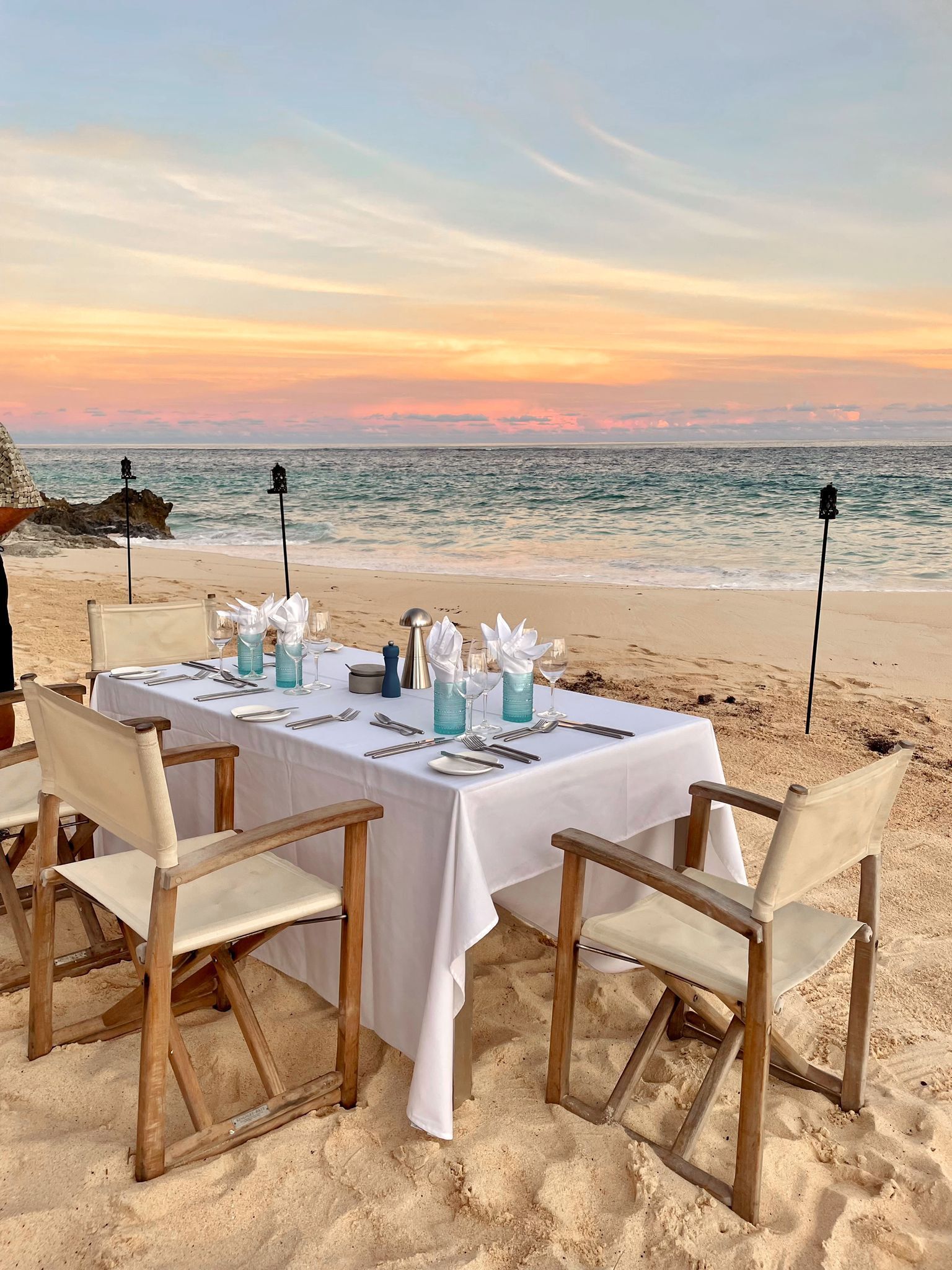 Mesa para dos personas en una playa arenosa, con vista al océano al atardecer.