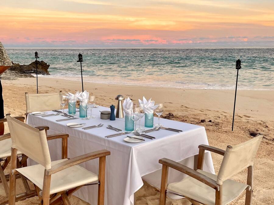 Mesa para dos personas en una playa arenosa, con vista al océano al atardecer.
