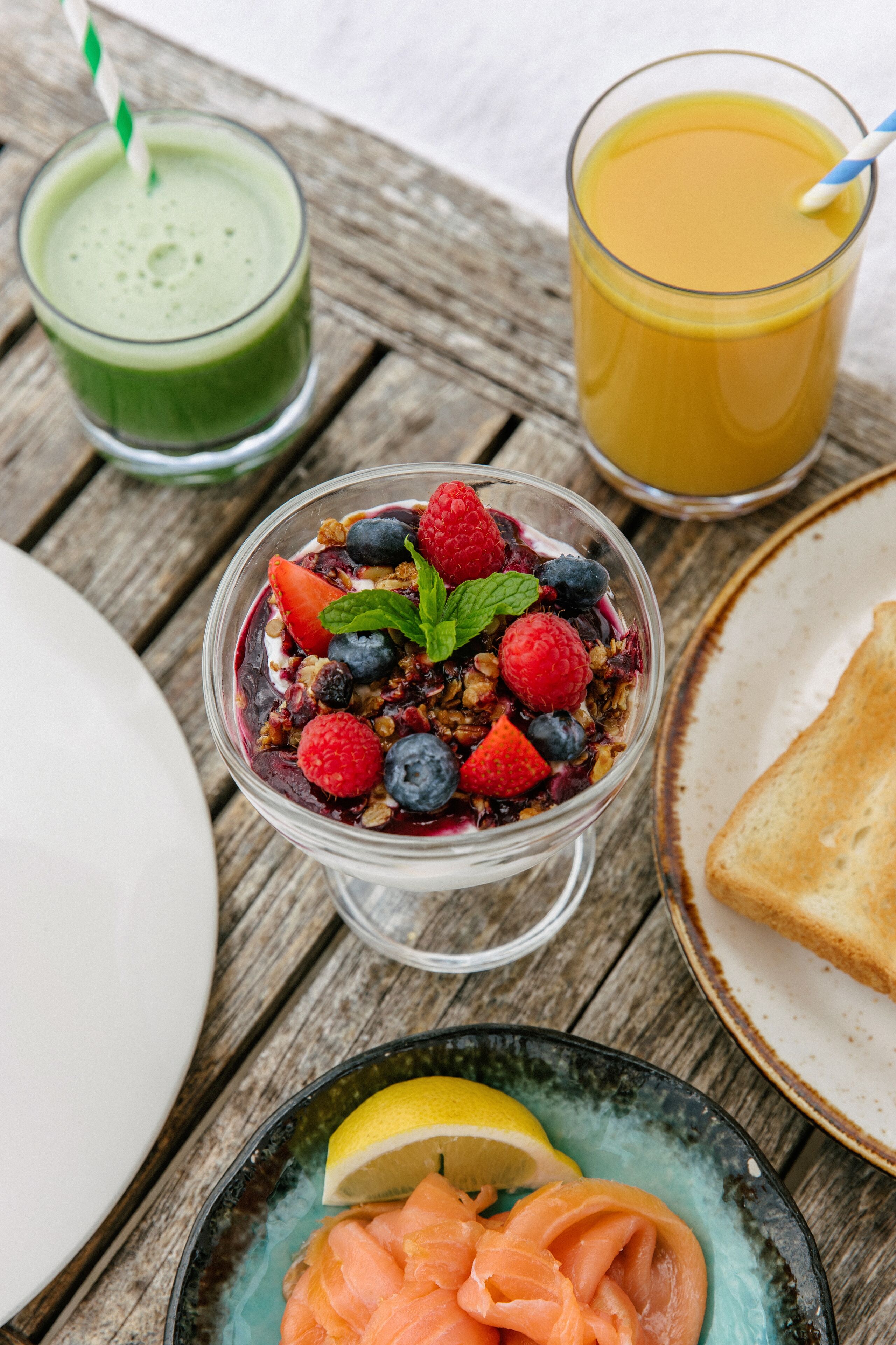 Uma mesa de café da manhã com um copo de smoothie verde, um copo de suco de laranja, uma tigela de iogurte com frutas vermelhas e granola, um prato de salmão defumado e uma fatia de pão torrado.