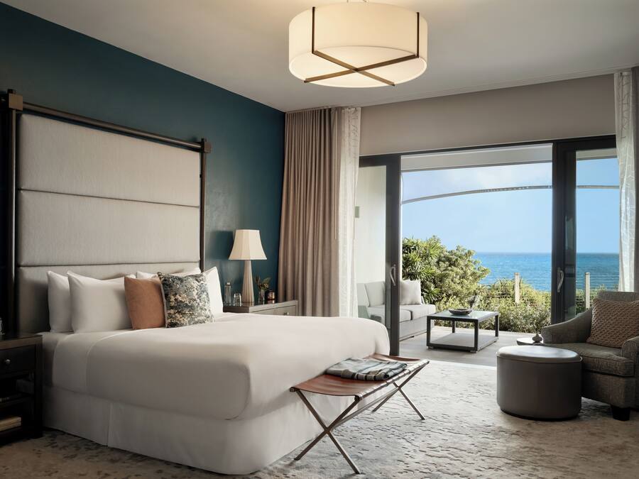 A modern hotel room with a large bed, a desk, a chair, and a view of the ocean.