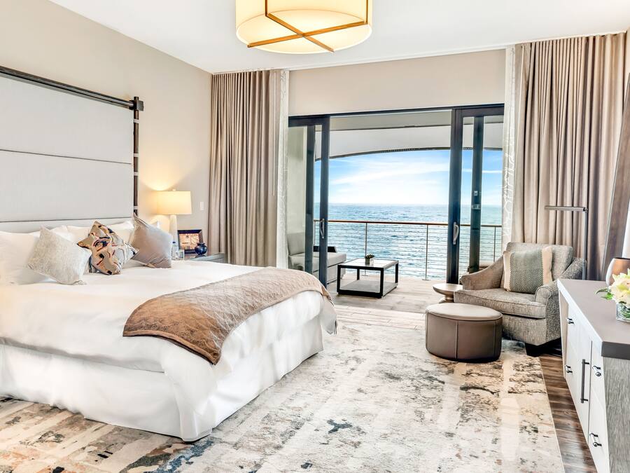 Quarto de hotel moderno com uma cama grande, área de estar com vista para o mar e varanda com cortinas.