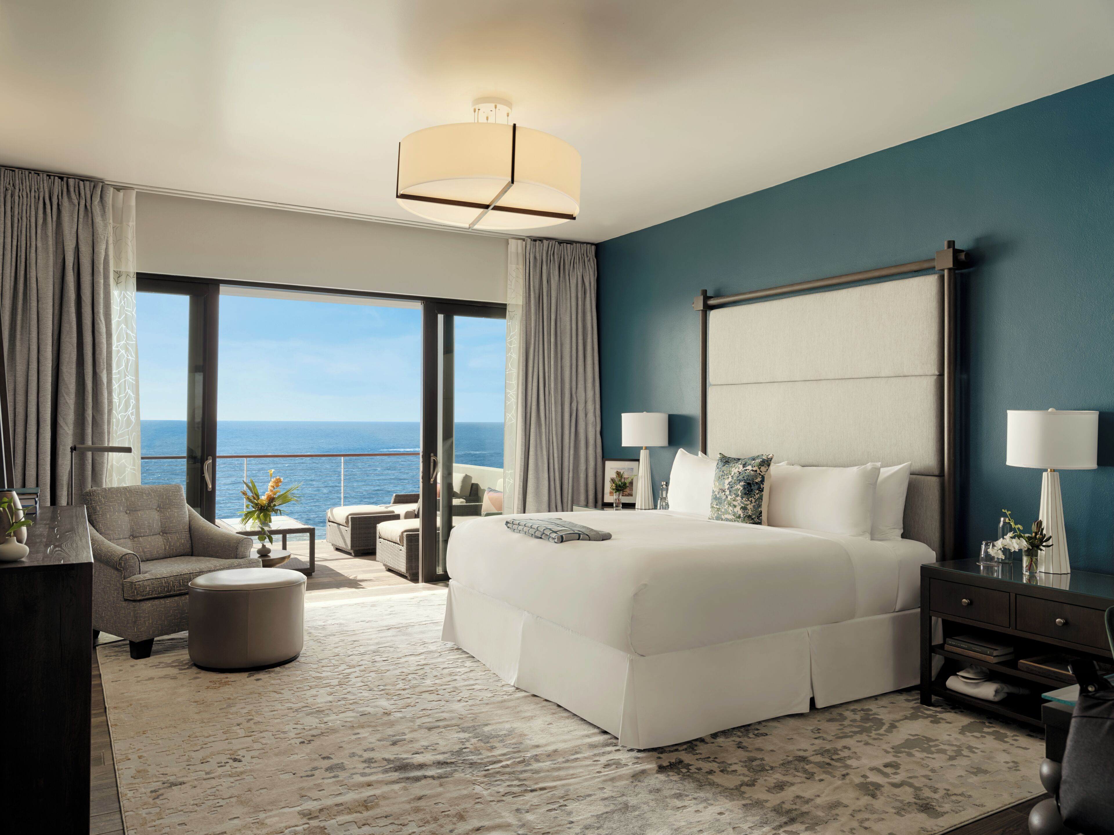 Quarto de hotel moderno com uma cama grande, área de estar, varanda vista para o mar e uma janela ampla.