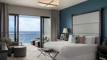 A modern hotel room with a large bed, a sofa, a small table, and a view of the sea.