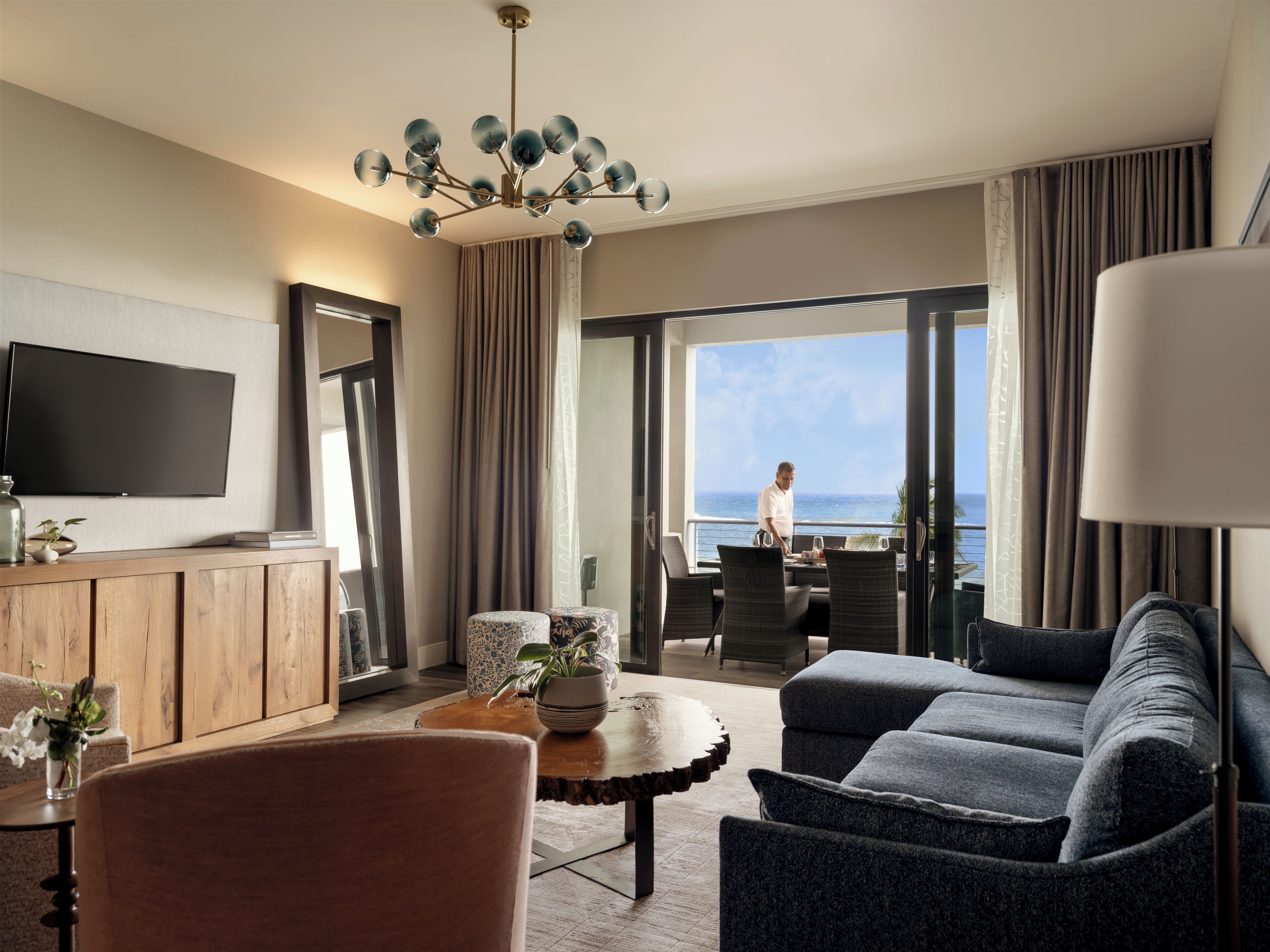 A modern hotel room with a large window, a flat-screen TV, a dining area, and a cozy seating area.